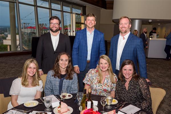 A group of seven well-dressed adults pose for a photo at a formal dinner event. Three individuals are standing behind a round table while four others are seated at the table. The table is set with glasses, plates of cake, salads, rolls, and bottles of wine and water. The background shows a large roo